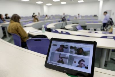 Se ve en primer plano una pantalla con gente conectada a una video llamada y difuminada atrás una sala de clases con estudiantes y profesor