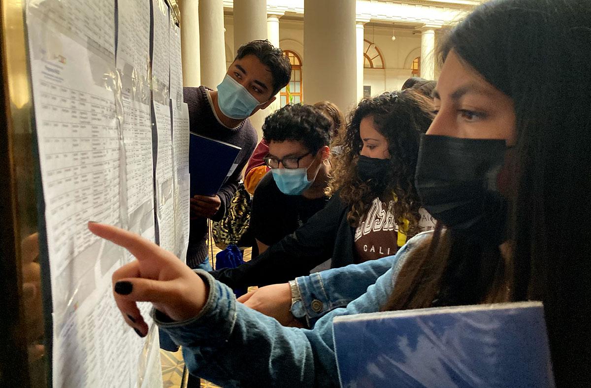 Estudiantes de la U. de Chile con mascarilla viendo unas listas en un mural
