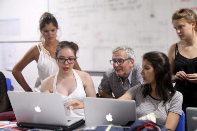 Cuatro estudiantes mujeres, observan algo en computadores junto a un profesor. De fondo, una pizarra.