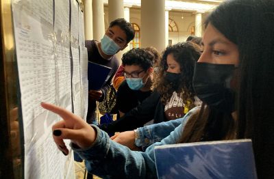 Estudiantes de la Universidad de Chile con mascarilla, revisando listas en un mural. Foto de Alejandra Fuenzalida